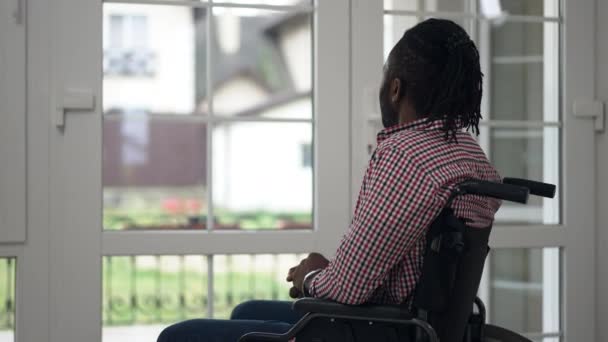 サイドビューの肖像画車いすのアフリカ系アメリカ人障害者の男性が自宅で窓の外を見てスローモーションで手を振っ。肯定的な自信に触発された麻痺した男屋内でリビングルーム挨拶隣人. — ストック動画