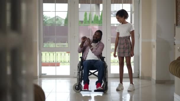 Wide shot portrait happy African American disabled man in wheelchair and teenage girl dancing in slow motion laughing gesturing thumbs up. Relaxed confident cheerful father and daughter in living room — Vídeo de Stock