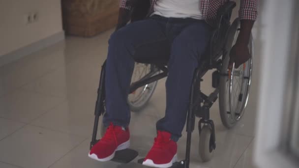 Uomo afroamericano irriconoscibile che rotola sulla sedia a rotelle al rallentatore sparando attraverso il vetro. Giovane disabile paralizzato in salotto o in casa di riposo. Concetto di vita assistita e disabilità. — Video Stock