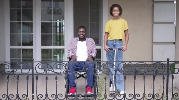 Retrato de feliz padre afroamericano discapacitado en silla de ruedas e hija adolescente con patinete scooter posando con sonrisa dentada y mirándose el uno al otro. Amplio tiro alegre hombre y chica en el porche. — Vídeo de stock