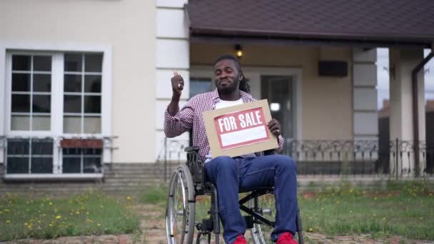 Afro-americano alegre confiante agente de vendas com deficiência em casa de publicidade em cadeira de rodas apontando para o fundo gesto polegar para cima sorrindo. Homem positivo vendendo imóveis posando em câmera lenta. — Vídeo de Stock