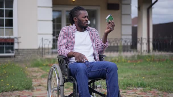 Retrato de un hombre afroamericano discapacitado en silla de ruedas al aire libre mirando la casa de juguetes con una adolescente borrosa saltando en cámara lenta al fondo en el porche. Pensativo tipo enfermo planeando la venta de casa. — Vídeo de stock