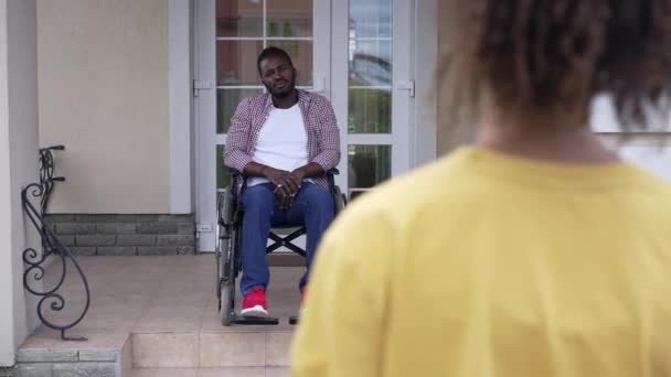 장애를 가진 아프리카 계 미국인 아버지가 휠체어를 타고 학교로 떠나는 10 대 딸에게 작별 인사를 하고 있습니다. 야외 현관 앞 마당에 있는, 자랑 스러운 부모가 소녀였을 때 모습. — 비디오