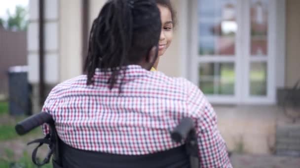 Happy teenage African American girl hugging disabled man in wheelchair smiling looking at camera. Portrait of cheerful daughter embracing father outdoors. Shooting over shoulder. — Stockvideo