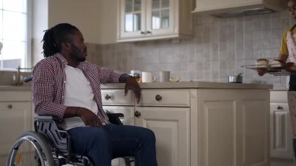 Positivo afroamericano discapacitado hombre en silla de ruedas hablando con una adolescente en delantal postre para hornear en la cocina. Retrato de padre feliz admirando a hija adolescente cocinando en casa sonriendo. — Vídeo de stock