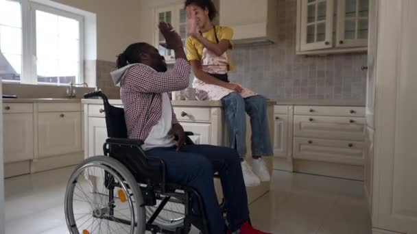 Geniş açılı mutlu, engelli, tekerlekli sandalyedeki Afrikalı Amerikalı adam ve gülümseyen genç kız yavaş çekimde el kol hareketi yaparak kameraya gülümsüyor. Kendine güvenen babanın portresi ve kızı mutfakta. — Stok video