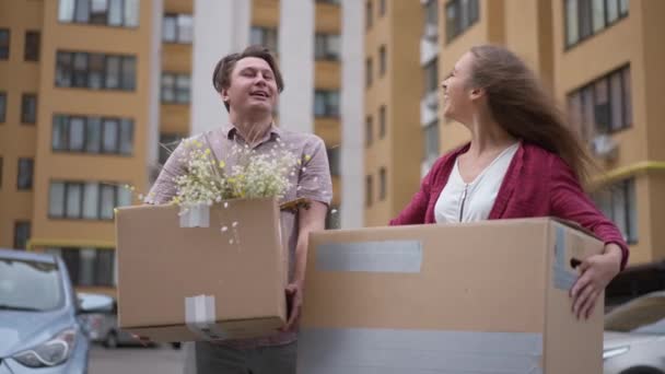 Excited young couple standing with boxes at parking lot admiring house and looking at camera smiling. Portrait of happy Caucasian husband and wife moving in new apartment posing in slow motion. — Vídeo de Stock