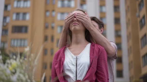 Young beautiful wife standing at new house with husband covering eyes with hands. Portrait of excited Caucasian woman outdoors and man surprising spouse with moving in new apartment. Slow motion. — Video Stock