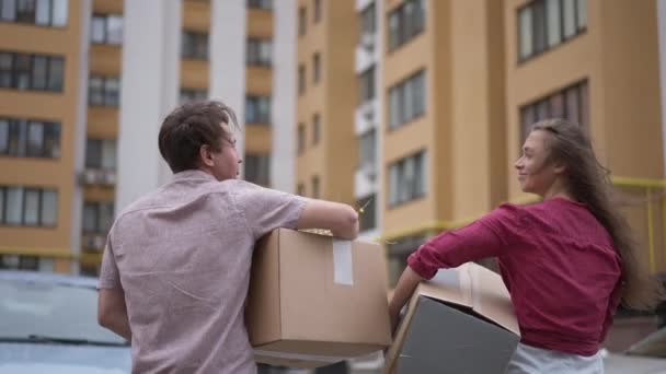 Tracking shot happy couple walking in slow motion on parking lot with cardboard boxes talking. Back view satisfied Caucasian man and woman moving in new apartment house. Tenancy and ownership concept. — Video Stock