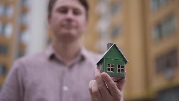 Close-up casa de juguete y llave en manos masculinas con borrosa feliz hombre caucásico sonriendo de fondo. Propietario del apartamento satisfecho de pie al aire libre en el edificio posando en cámara lenta. — Vídeos de Stock