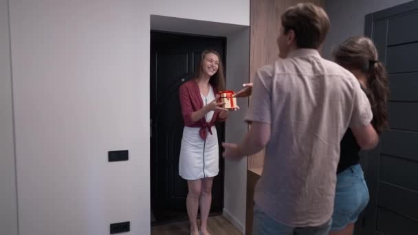 Happy couple standing in apartment with friend entering smiling holding gift talking in slow motion. Caucasian husband and wife welcoming guest in new flat indoors. Rent and tenancy. — Stock Video