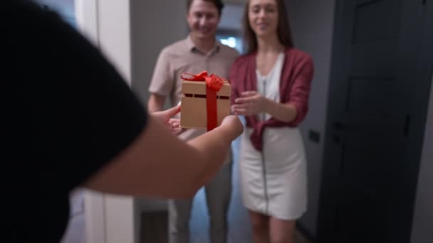 Rack focus from gift box in female hands to apartment owners smiling greeting guest indoors in slow motion. Positive loving Caucasian couple meeting friend in new flat. Real estate mortgage and rent. — Vídeo de Stock