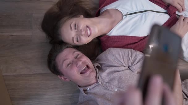 Suami dan istri kaukasia yang bersusah payah mengambil foto selfie di smartphone berbaring di lantai parket. Pasangan serampangan bersenang-senang di dalam rumah pada akhir pekan. Relaxation dan gaya hidup. — Stok Video
