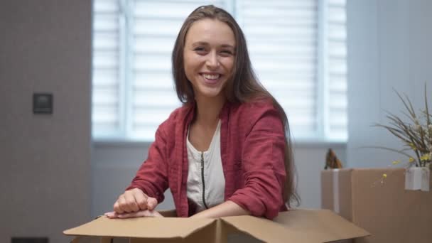 Beautiful happy woman gesturing thumbs up smiling looking at camera sitting in box indoors. Portrait of excited Caucasian millennial lady moving in new apartment posing in slow motion. — стокове відео