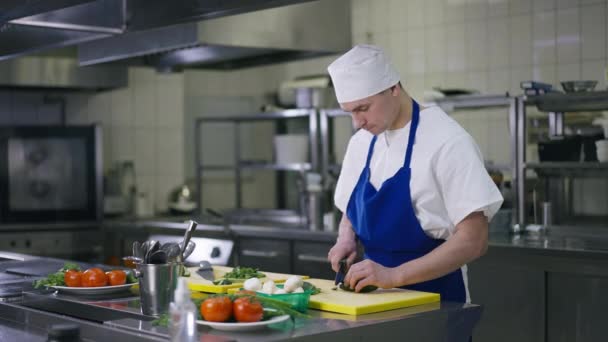 Kavkazský kuchař krájí okurku jíst plátek jako kuchař procházející v pozadí. Portrét muže v uniformě pracující v komerční kuchyni v restauraci. Koncepce zaměstnání a povolání. — Stock video