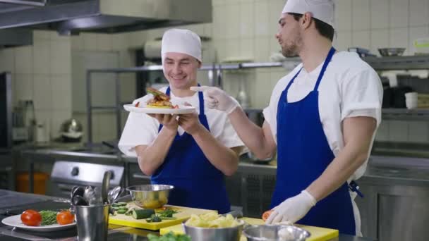 Smiling cook boasting served seafood on plate to chef in kitchen indoors. Positive Caucasian professional man showing dish as colleague talking admiring meal. Professionalism concept. — Video Stock
