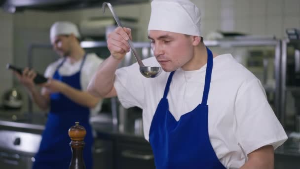 Caucasian chef tasting soup adding salt to dish standing in kitchen with cook preparing food at background. Portrait of confident man dressing meal in restaurant. Profession and taste concept. — Video Stock