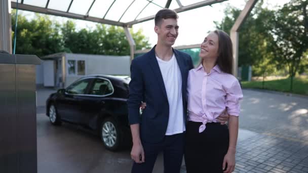 Satisfied Caucasian couple gesturing thumbs up smiling looking at camera posing at car wash station outdoors. Confident smiling man and woman hugging posing in slow motion. Auto industry. — Vídeo de Stock