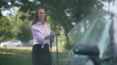 Slim attractive charming woman cleaning automobile with high pressure washer at self service station outdoors. Portrait of smiling beautiful Caucasian lady washing vehicle on sunny day.