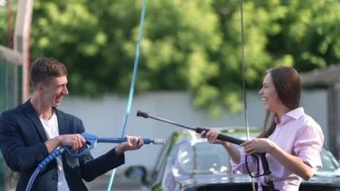 Joyful couple imitating shooting with high pressure washer in slow motion laughing having fun at car wash service. Happy loving Caucasian man and woman enjoying cleaning automobile outdoors.