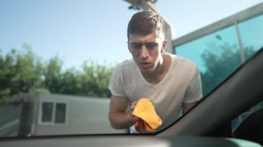 Professional Caucasian man cleaning vehicle windshield in slow motion in sunrays outdoors. Portrait of concentrated male employee washing automobile at car wash service. Auto maintenance concept.