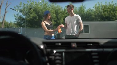Young Caucasian man passing cloth to woman cleaning vehicle at car wash service outdoors in sunshine. Positive male and female employees washing automobile polishing surface in slow motion.