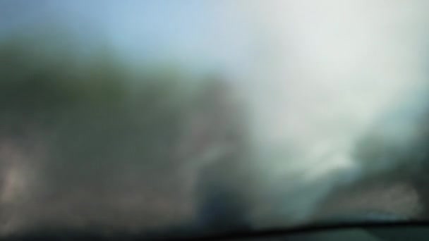Крупним планом автомобільне лобове скло з водяним розпиленням з мийкою високого тиску і розмитою усміхненою молодою жінкою, що працює на фоні. Зйомки зсередини автомобіля прибирання автомобіля в автомийці . — стокове відео