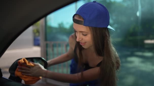 Positive brunette woman cleaning side view mirror in slow motion outdoors in sunshine. Portrait of Caucasian employee washing automobile at car wash service. — Wideo stockowe