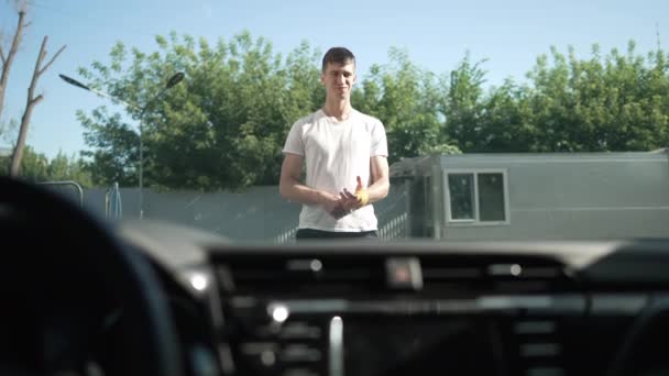 Satisfied Caucasian young man admiring clean automobile at car wash service outdoors. Portrait of smiling confident employee standing in sunshine looking at vehicle. Shooting from inside car. — Stockvideo