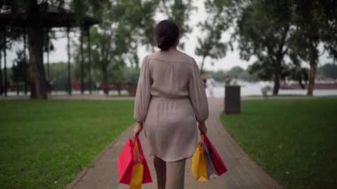Live camera follows confident senior woman walking with shopping bags outdoors. Back view elegant stylish Caucasian retiree strolling in park on Black Friday sales season. Fashion and lifestyle.