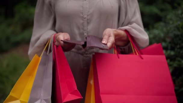 Unrecognizable Caucasian senior woman opening empty wallet showing no money inside. Wrinkled female hands with shopping bags and billfold standing outdoors in park. Spending and sales concept. — Wideo stockowe