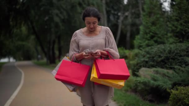 Wealthy satisfied senior Caucasian woman with shopping bags counting cash money walking away leaving in park outdoors. Portrait of happy retiree strolling with purchases on Black Friday smiling. — Stock video