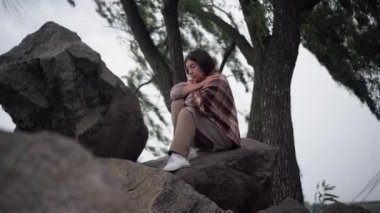 Depressed sad senior woman sitting on rocks with wind blowing tree branches at background. Wide shot portrait of beautiful frustrated Caucasian retiree procrastinating outdoors thinking looking away.