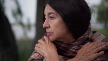 Close-up senior woman with black long hair and brown eyes looking away sitting in park on windy day. Portrait of thoughtful unhappy Caucasian retiree alone on cloudy day outdoors.