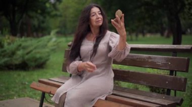 Elegant beautiful senior woman sitting on bench in city park admiring reflection in hand mirror smiling adjusting hair. Portrait of confident gorgeous slim Caucasian retiree resting outdoors.