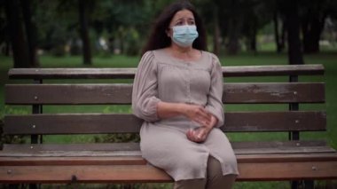 Front view portrait stressed Caucasian senior woman in Covid-19 face mask coughing sitting on bench in park outdoors. Worried frightened retiree with symptoms of coronavirus illness looking around.