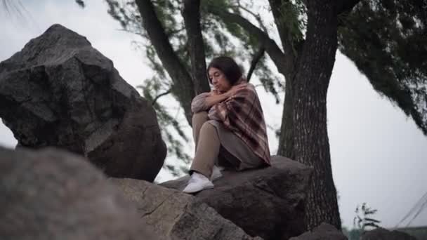 Depressed sad senior woman sitting on rocks with wind blowing tree branches at background. Wide shot portrait of beautiful frustrated Caucasian retiree procrastinating outdoors thinking looking away. — Stock video