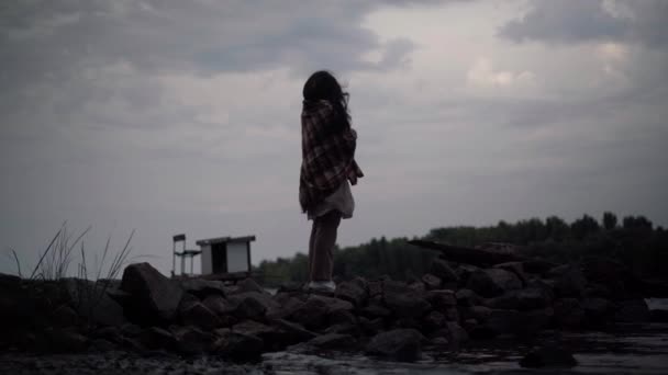 Silhouette portrait of senior Caucasian woman standing on rocky river bank on overcast evening in twilight looking away. Wide shot thoughtful sad retiree procrastinating in dusk. Aging and loneliness. — Stock video