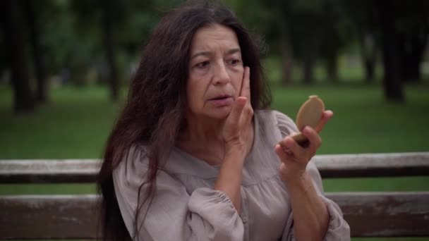 Worried sad senior woman looking in hand mirror touching wrinkled face sighing looking away. Portrait of anxious Caucasian retiree dissatisfied with faded beauty thinking sitting in park outdoors. — Stockvideo