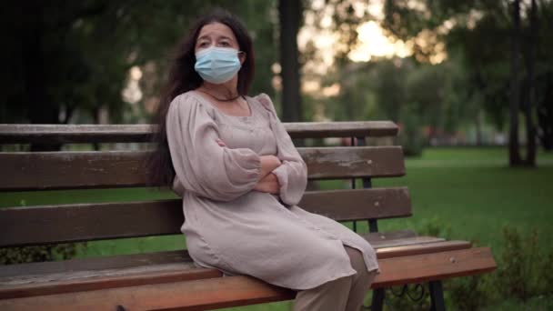 Lonely sad senior woman in Covid face mask sitting on bench in park sighing. Portrait of upset Caucasian retiree alone outdoors on coronavirus pandemic outbreak. New normal concept. — стоковое видео