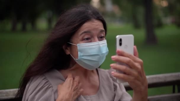 Positive beautiful senior woman in Covid-19 face mask waving talking smiling using video chat app on smartphone. Portrait of happy Caucasian retiree messaging online resting in park on pandemic. — Stockvideo