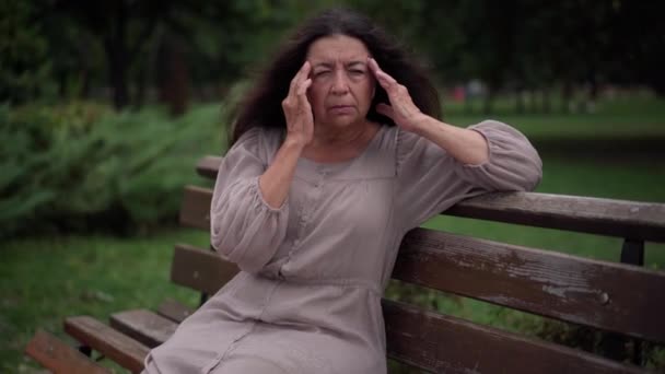 Ill tired sad senior woman rubbing temples sitting on bench in park. Portrait of Caucasian retiree with headache migraine outdoors on windy overcast day. Chronic illness and lifestyle concept. — Video Stock