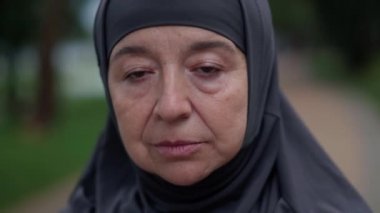 Close-up face sad senior Muslim woman in hijab looking at camera with brown eyes standing outdoors. Headshot portrait of upset Middle Eastern retiree posing in park. Aging and lifestyle concept.