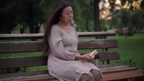 Portrait of frustrated senior Caucasian woman eating sandwich sitting on bench in park on windy overcast day. Medium shot lonely upset retiree thinking biting chewing junk food outdoors. — Stok video
