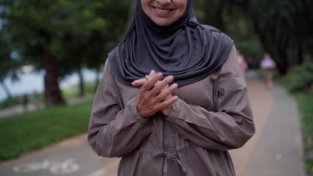 Unrecognizable smiling senior woman in hijab putting hands together standing on park alley. Positive Middle Eastern retiree enjoying leisure outdoors on spring summer day. Confidence and lifestyle. — Vídeo de Stock