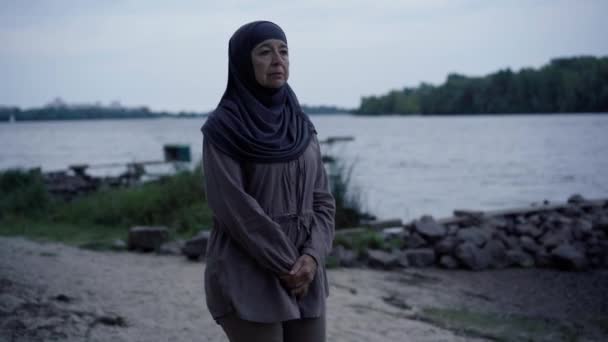 Sad senior woman in hijab walking on river bank looking away thinking. Portrait of depressed Middle Eastern Muslim retiree strolling outdoors on cloudy autumn day. Aging and sadness concept. — Stockvideo