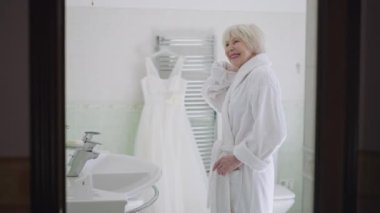 Happy senior Caucasian bride in bathrobe standing in bathroom admiring reflection in mirror and wedding dress hanging at background. Side view beautiful woman with grey hair getting ready for marriage