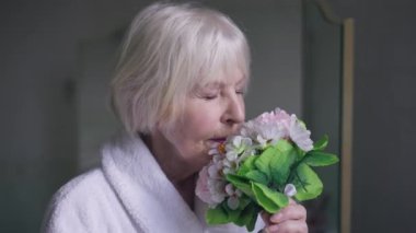 Close-up portrait of beautiful senior woman smelling bridal bouquet drinking champagne looking out the window in the morning. Thoughtful Caucasian bride with grey hair indoors at home. Natural beauty.
