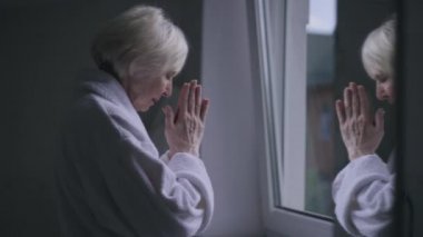 Side view reflection in mirror of sad senior Caucasian woman praying standing at window indoors. Upset depressed lonely retiree in bathrobe with hands together asking God for help and thanking.