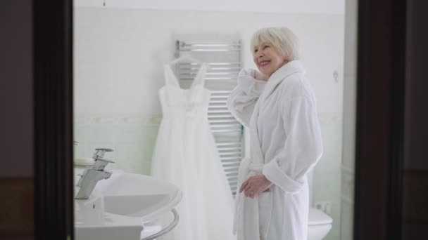 Happy senior Caucasian bride in bathrobe standing in bathroom admiring reflection in mirror and wedding dress hanging at background. Side view beautiful woman with grey hair getting ready for marriage — Stockvideo
